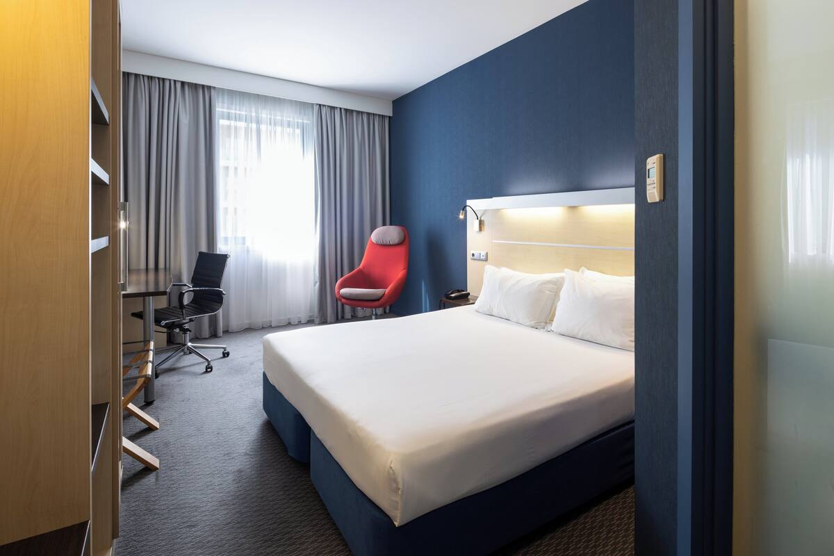 A hotel room with a large bed, a red chair, a desk, and a window with curtains.