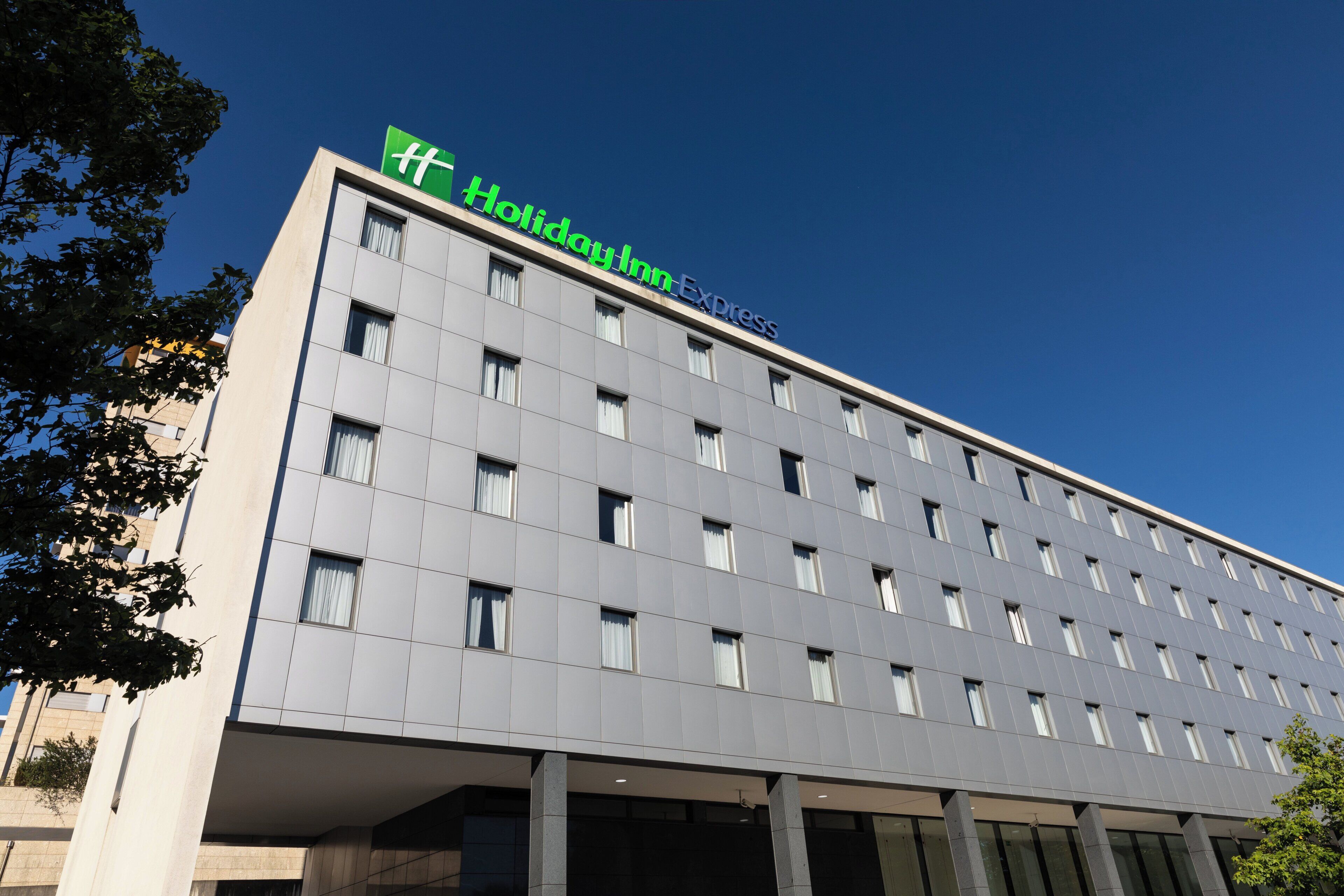 A modern hotel building with a flat facade, multiple windows, and a prominent green sign indicating it is a Holiday Inn Express.