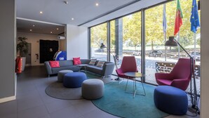 A modern lobby with a grey sofa, red and blue chairs, and a round table. There are large windows with flags and a view of the outside. - Holiday Inn Express Porto - Exponor by IHG (Matosinhos)