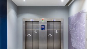 A pair of elevator doors with directional arrows, a blue emergency exit sign, and a map on the adjacent wall. - Holiday Inn Express Porto - Exponor by IHG (Matosinhos)