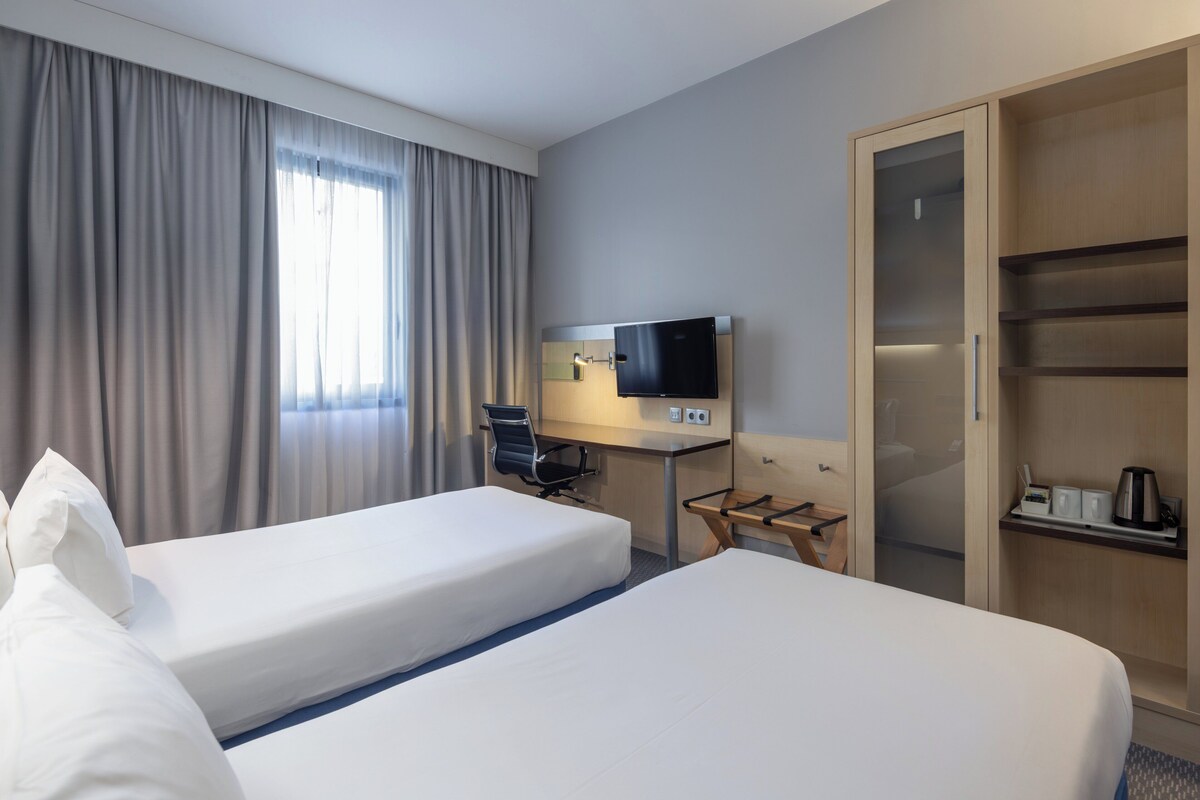 Ein Hotelzimmer mit zwei Betten, einem Schreibtisch mit Fernseher, einem Kleiderschrank und einem Fenster mit Vorhängen.