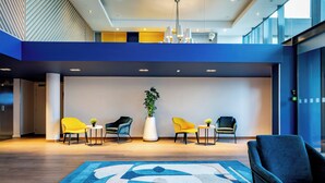 Un hall d’entrée moderne avec un tapis aux motifs bleus et blancs, un espace détente avec des fauteuils jaunes et bleus, et une grande plante en pot.