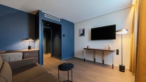 A modern living room with a sofa, a coffee table, a TV mounted on the wall, a desk, and a lamp. - Holiday Inn Mulhouse by IHG (Mulhouse)