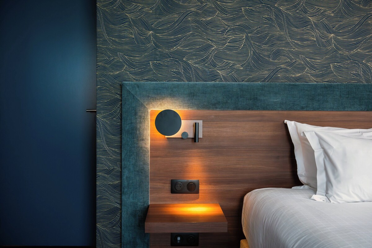 A hotel room with a wooden headboard, a bedside table with a lamp, and a patterned wall.