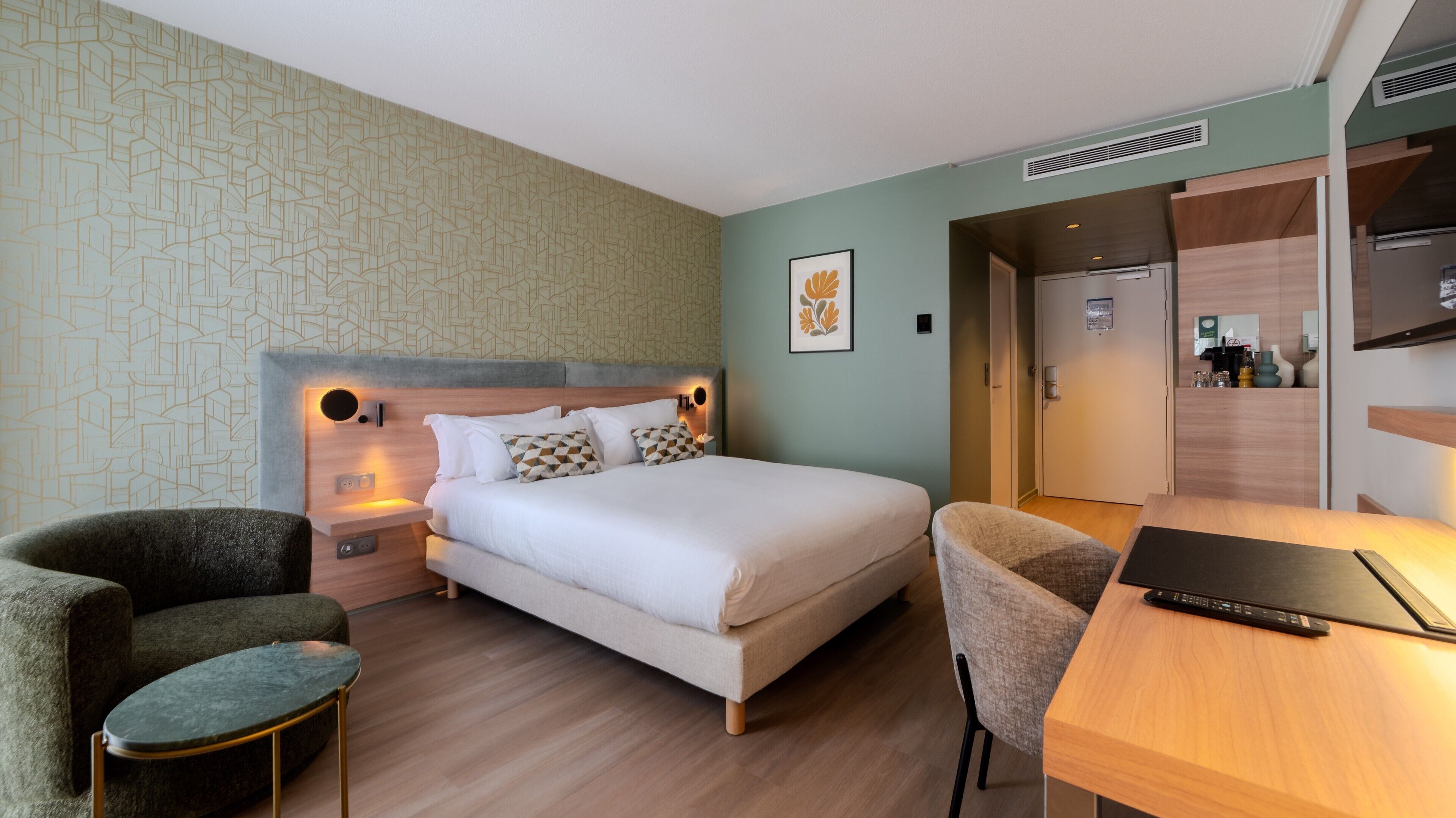 Une chambre d’hôtel comprenant un lit, deux fauteuils, un bureau avec un ordinateur portable, un mur orné d’un motif géométrique et une porte donnant sur une salle de bain.