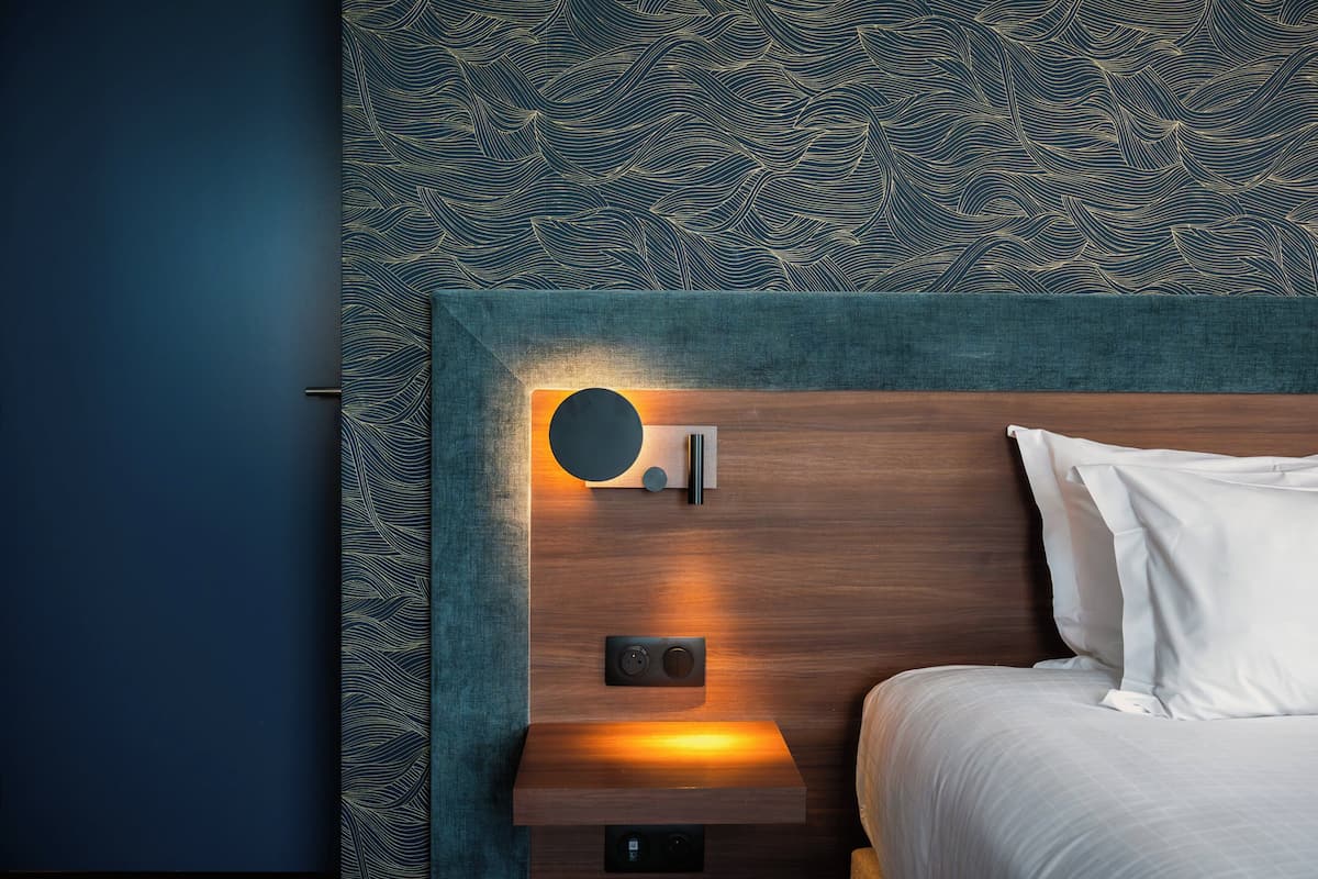 A hotel room with a wooden headboard, a bedside table with a lamp, and a patterned wall.