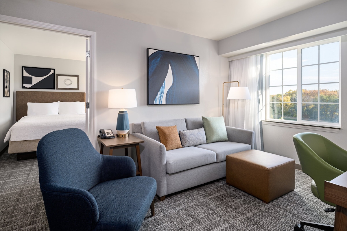 A hotel room with a bed, a grey sofa, a blue armchair, a green chair, and a window with a view of trees.