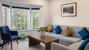 A modern living room with a grey sofa, blue armchair, and a coffee table. There are large windows with a view of trees and a framed picture on the wall.