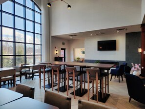 A modern dining area with high ceilings, large windows, and a flat-screen TV mounted on the wall.