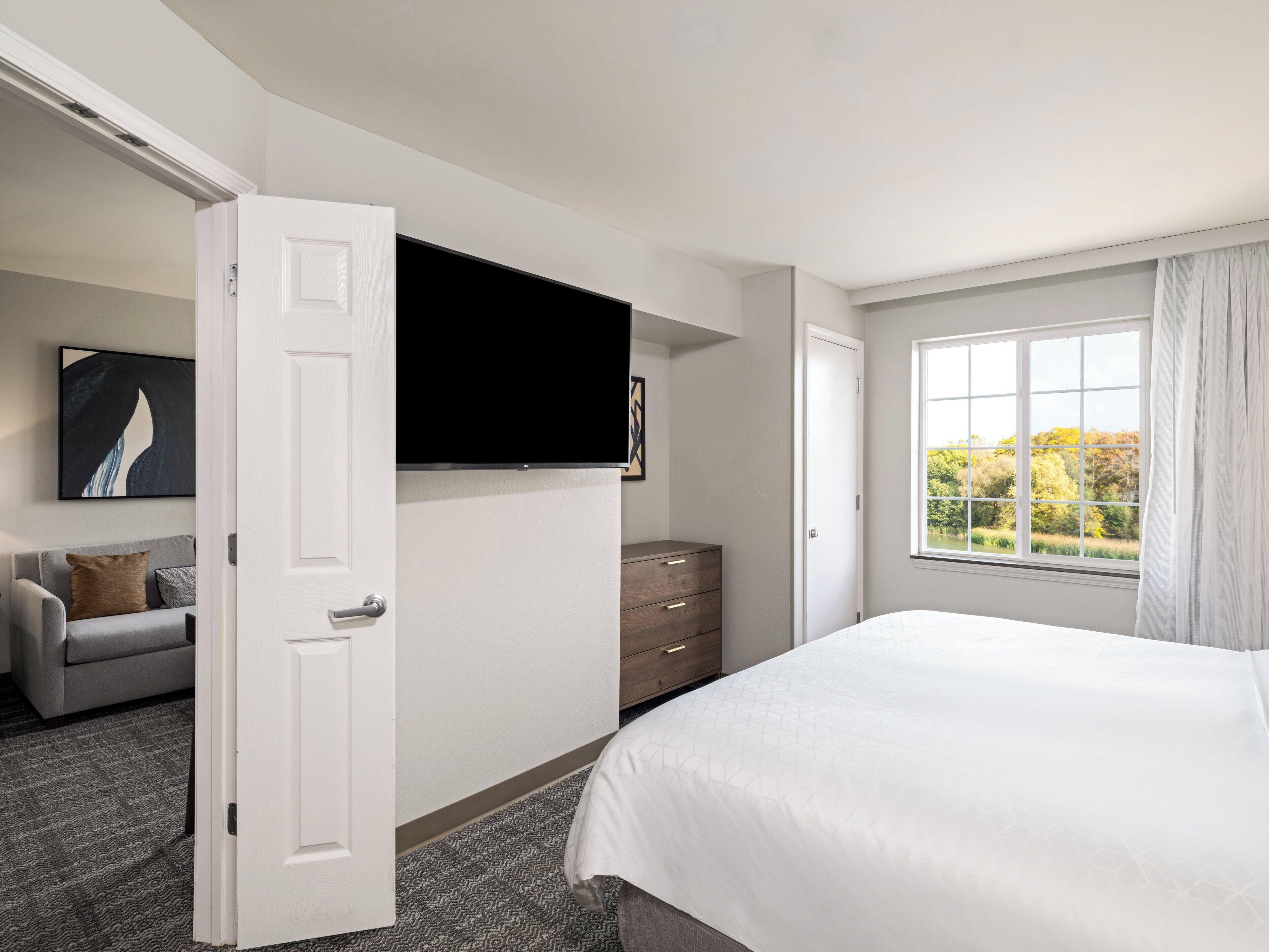 Une chambre d’hôtel avec un lit, un téléviseur fixé au mur, un canapé gris, une commode en bois et une fenêtre donnant sur des arbres.