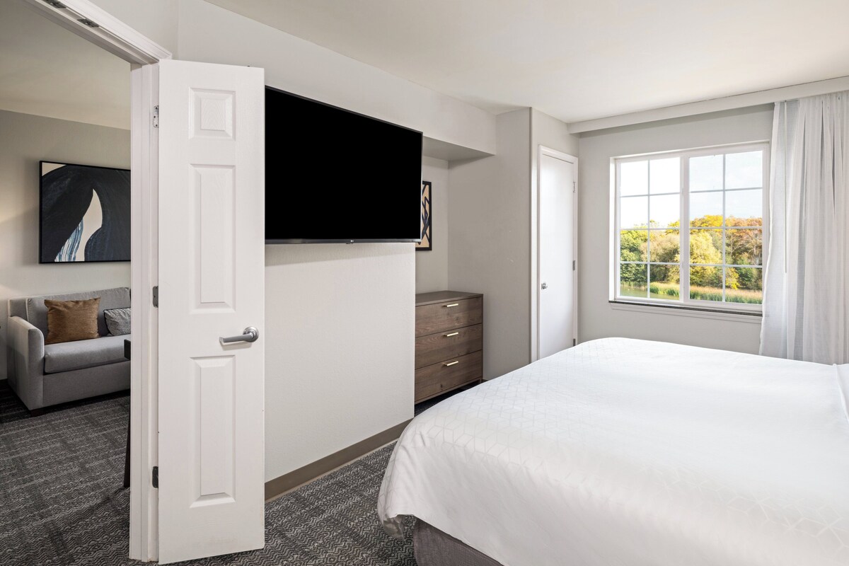 Een hotelkamer met een groot bed, een flatscreen televisie aan de muur, een grijze bank, een houten dressoir en een raam met uitzicht op bomen.