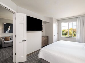 A hotel room with a bed, a TV mounted on the wall, a grey sofa, a wooden dresser, and a window with a view of trees. - Staybridge Suites Rochester University by IHG (Rochester)