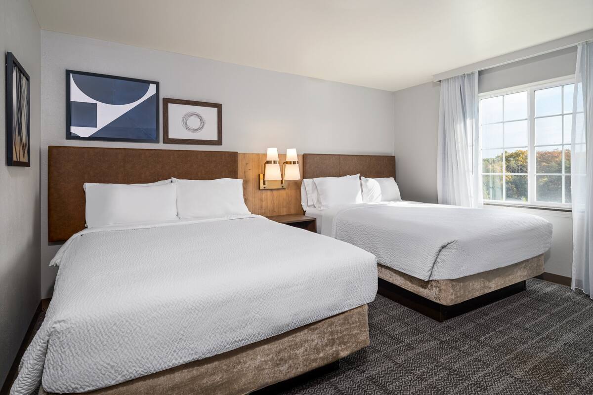 Quarto de hotel com duas camas, uma janela grande com cortinas e obras de arte nas paredes.