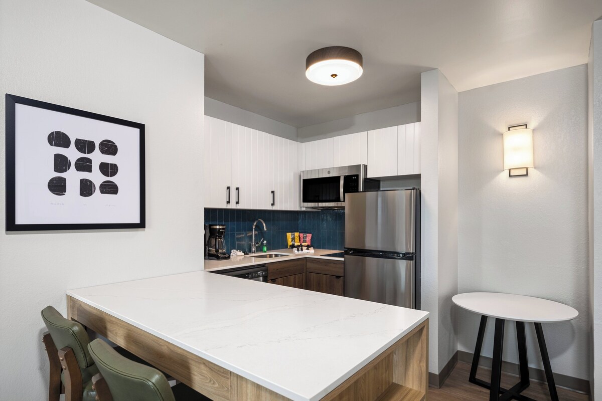 Een moderne keuken met witte kasten, een roestvrijstalen koelkast, een magnetron en een koffiezetapparaat. Er hangt een lijst met kunst aan de muur en er staat een klein, rond tafeltje met een kruk.