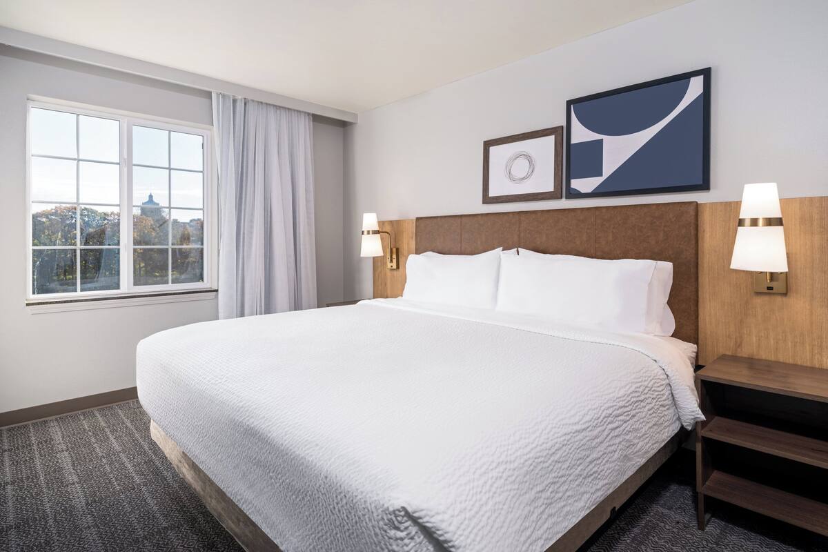 Quarto de hotel com uma cama grande, janela com cortinas, duas luminárias fixadas na parede e duas imagens emolduradas.