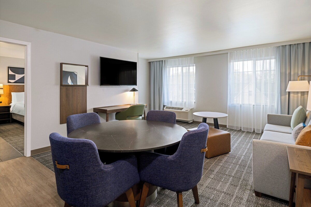 Een hotelkamer met een ronde tafel, twee blauwe stoelen, een bank, een aan de muur bevestigde televisie en een raam met gordijnen.