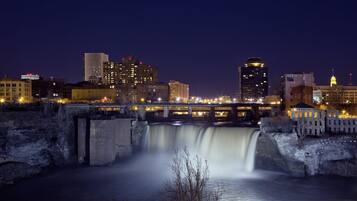 A nighttime cityscape with a bridge over a waterfall, illuminated buildings, and a river.