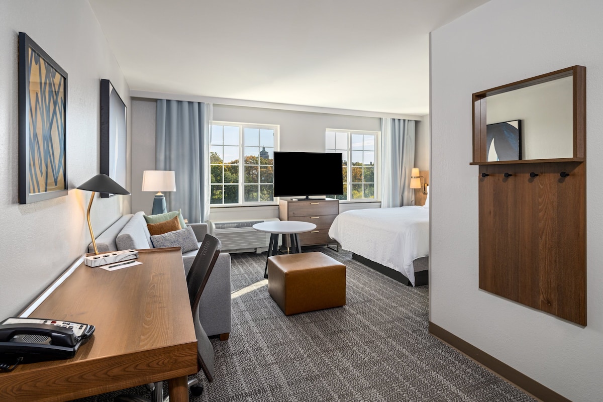 A hotel room with a bed, a desk with a telephone, a television, and a seating area with a chair and ottoman.