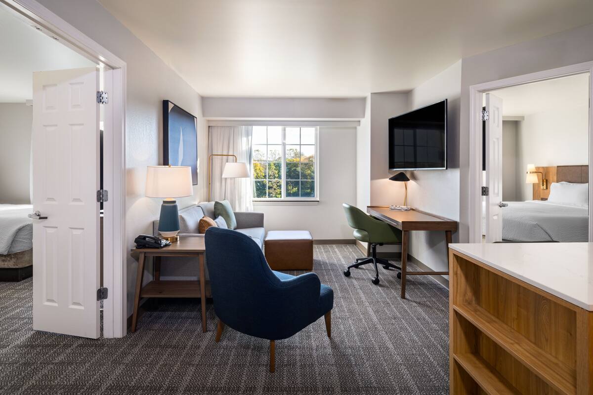 Quarto de hotel com cama, escrivaninha com cadeira, televisão e janela com vista para árvores.