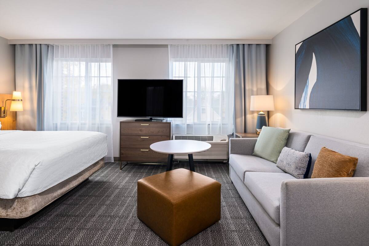 Quarto de hotel com cama, sofá, mesa de centro, televisão e um quadro na parede.