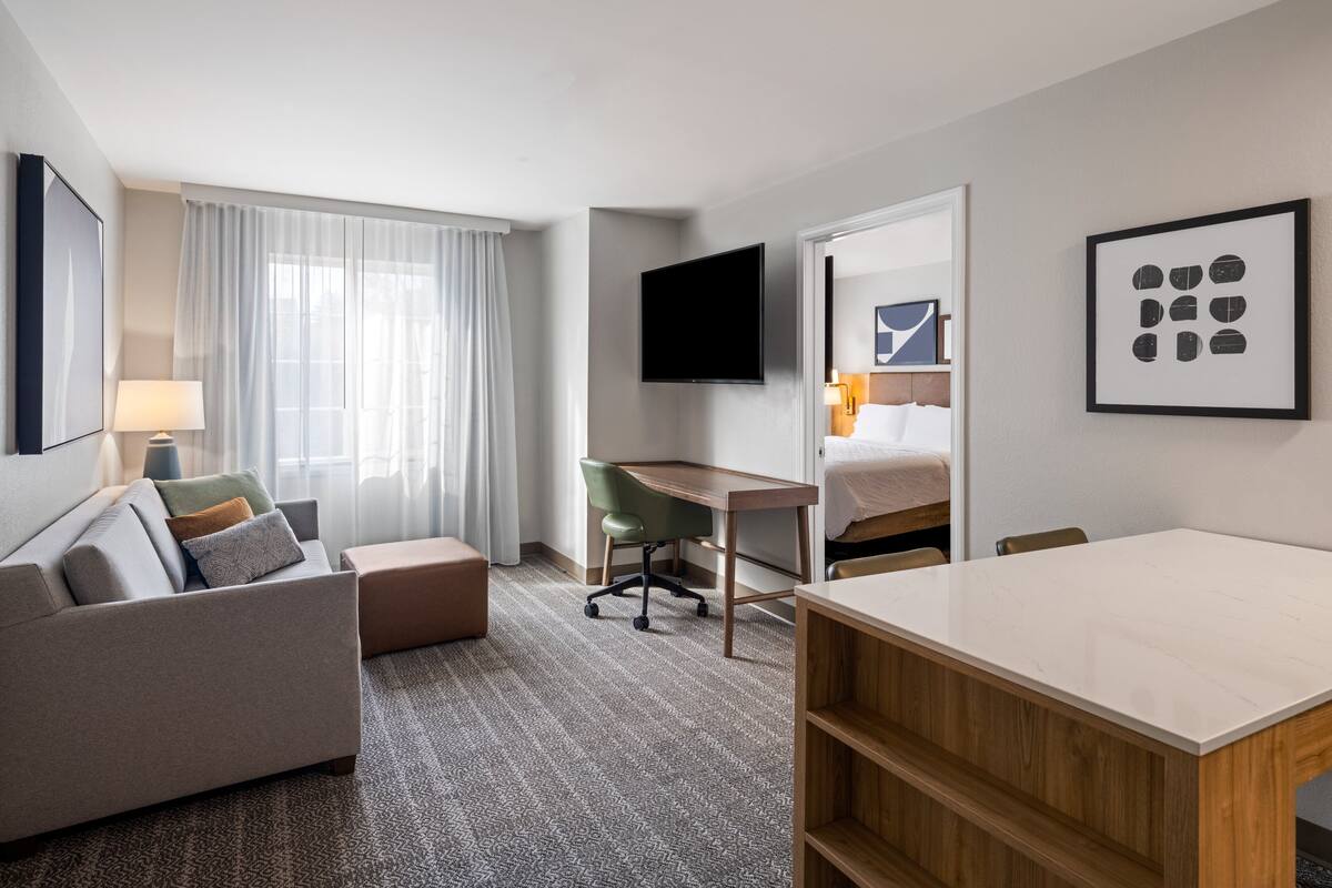 Quarto de hotel com sofá, escrivaninha, cadeira, cama, televisão e obras de arte nas paredes.