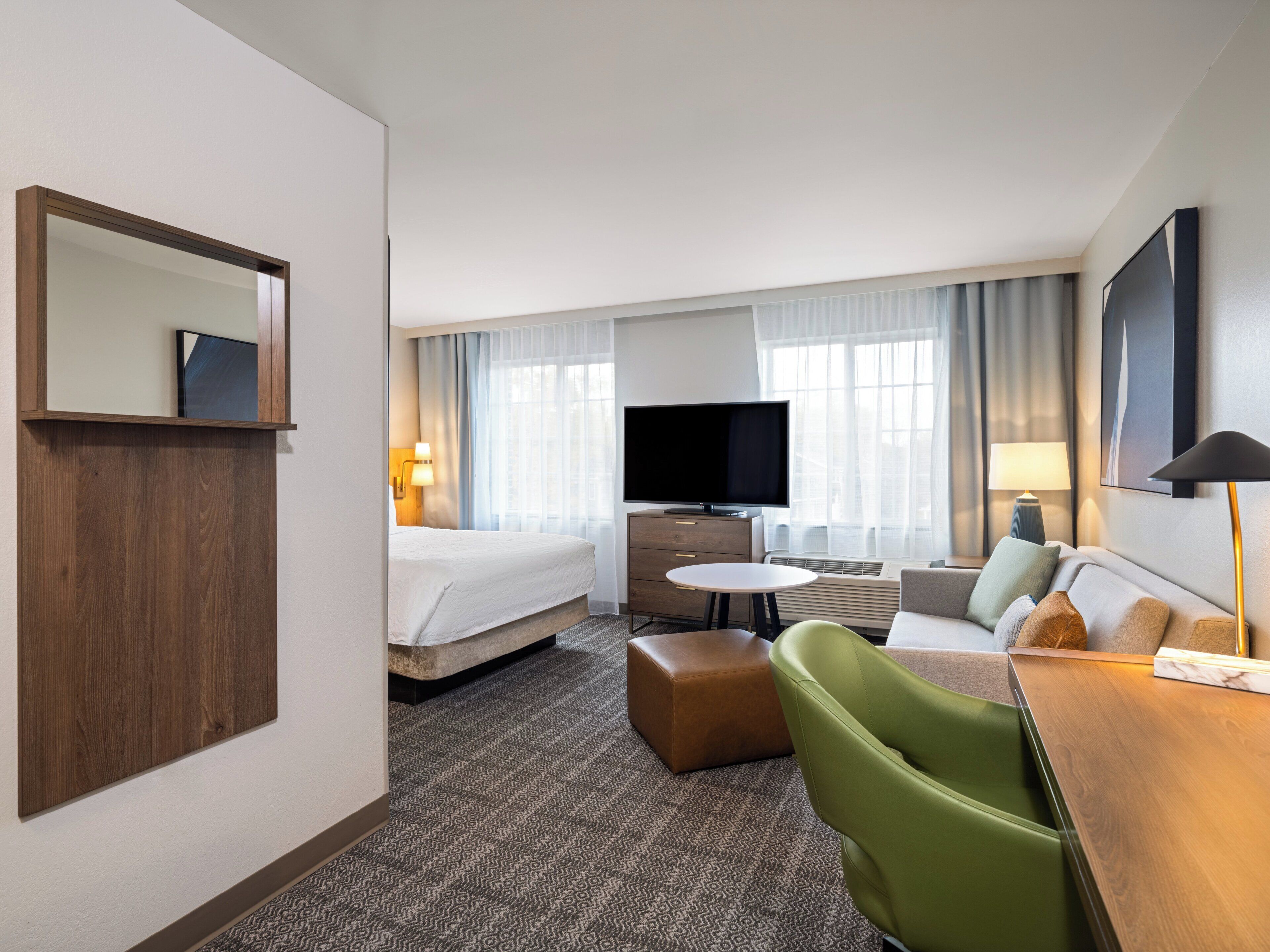 Une chambre d’hôtel comprenant un lit, une télévision, un bureau avec une chaise, un canapé et une grande fenêtre avec des rideaux.