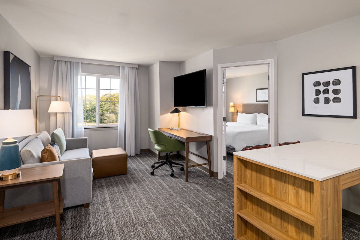 A hotel room with a bed, a desk, a chair, a sofa, a TV, and a window with curtains.