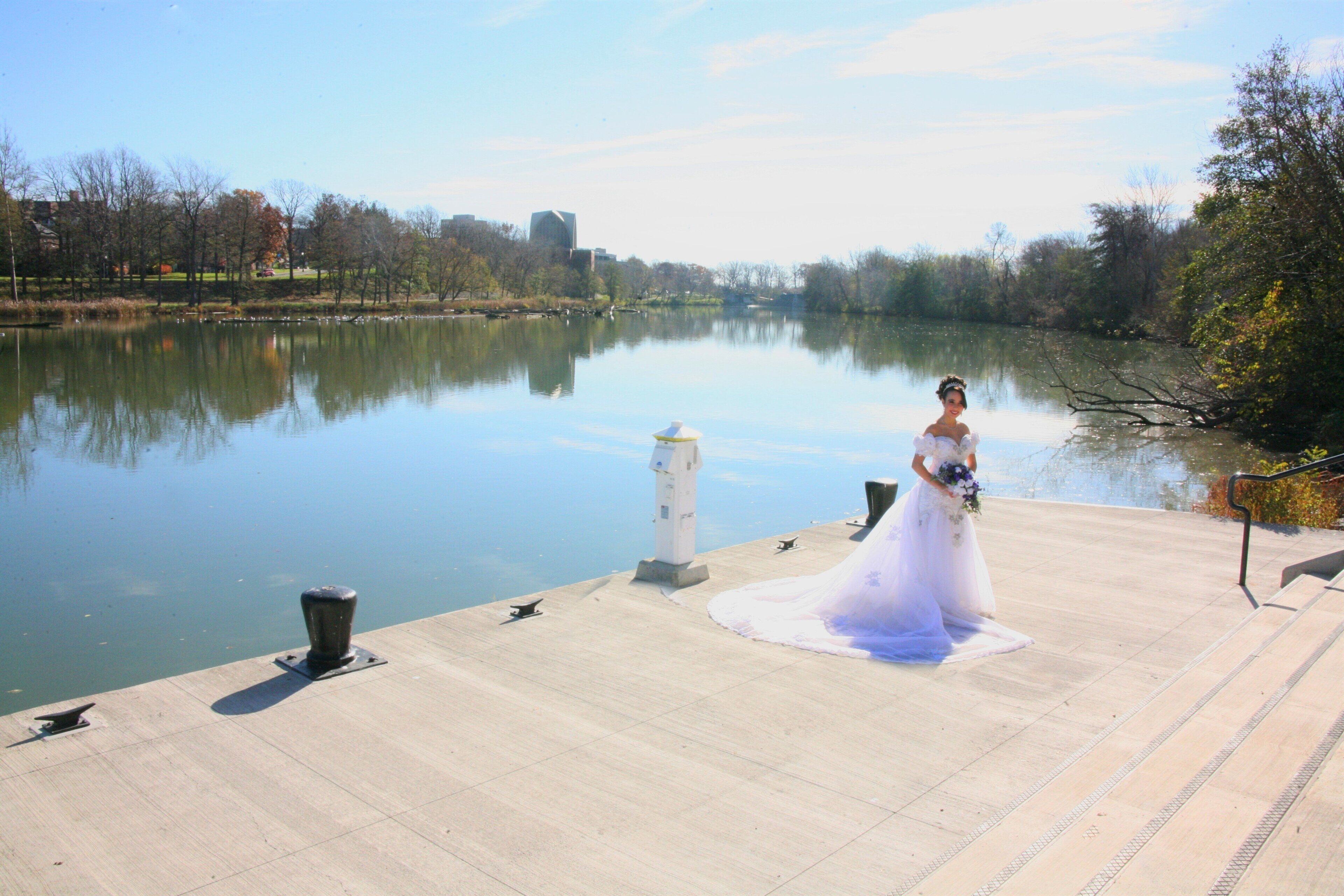 Eine Braut im Brautkleid steht an einem Steg an einem ruhigen Fluss, im Hintergrund Bäume und Gebäude.