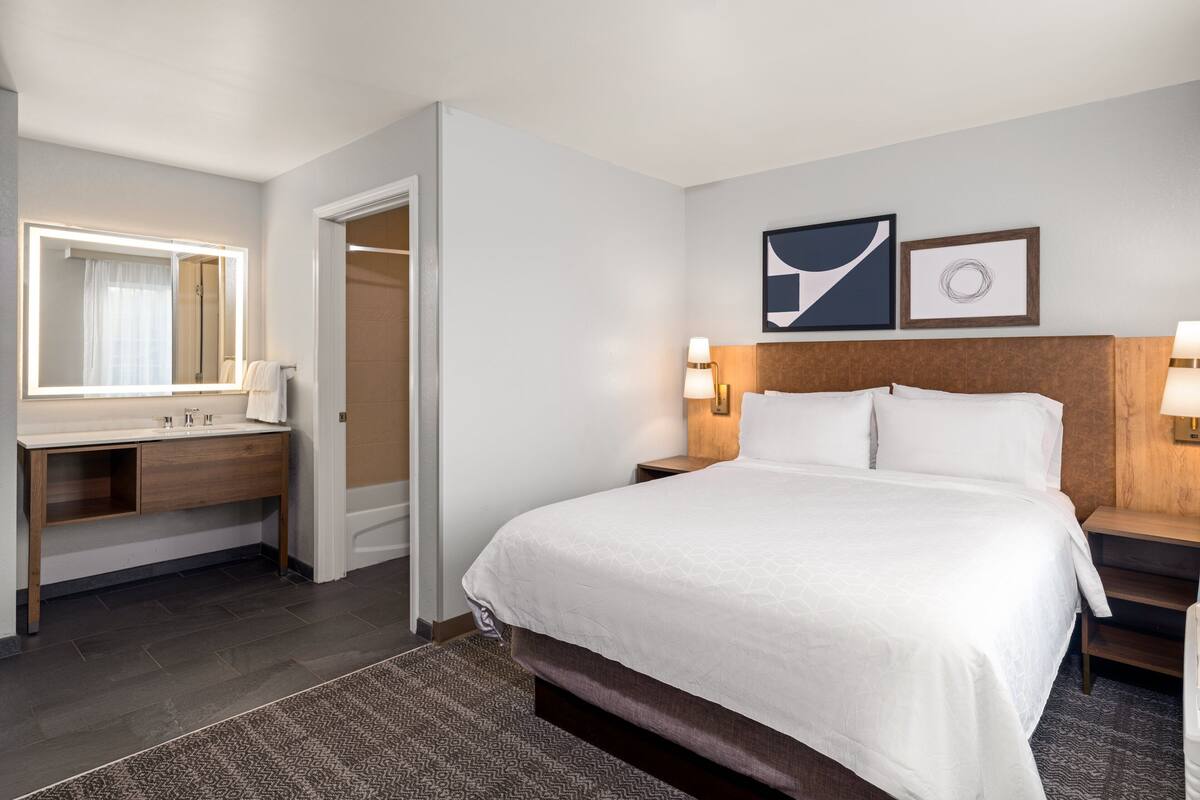 Quarto de hotel com uma cama grande, uma mesinha de cabeceira com abajur, um banheiro com espelho e chuveiro.