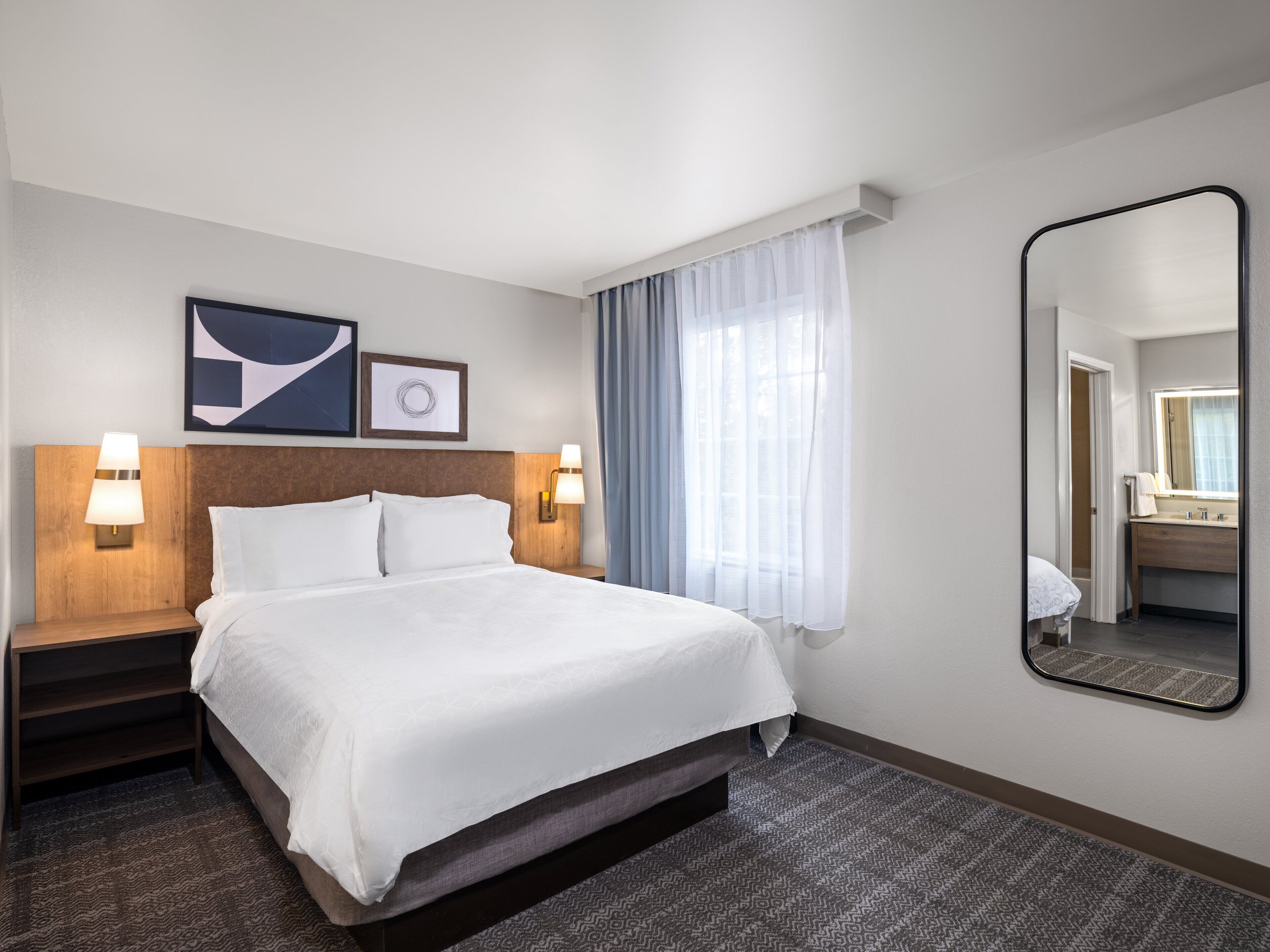 Habitación de hotel con cama, dos lámparas de noche, un espejo grande y una ventana con cortinas.