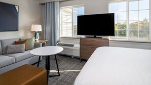 A hotel room with a large bed, a sofa, a small table, a TV, and a window with a view of trees and a building.