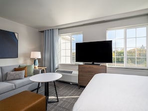 A hotel room with a large bed, a sofa, a small table, a TV, and a window with a view of trees and a building.