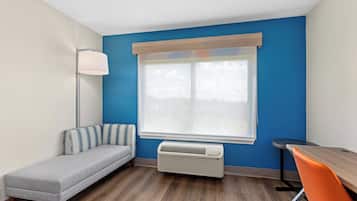 A room with a blue wall, a window with a white blind, a gray sofa, a standing lamp, a wooden table, and an orange chair.