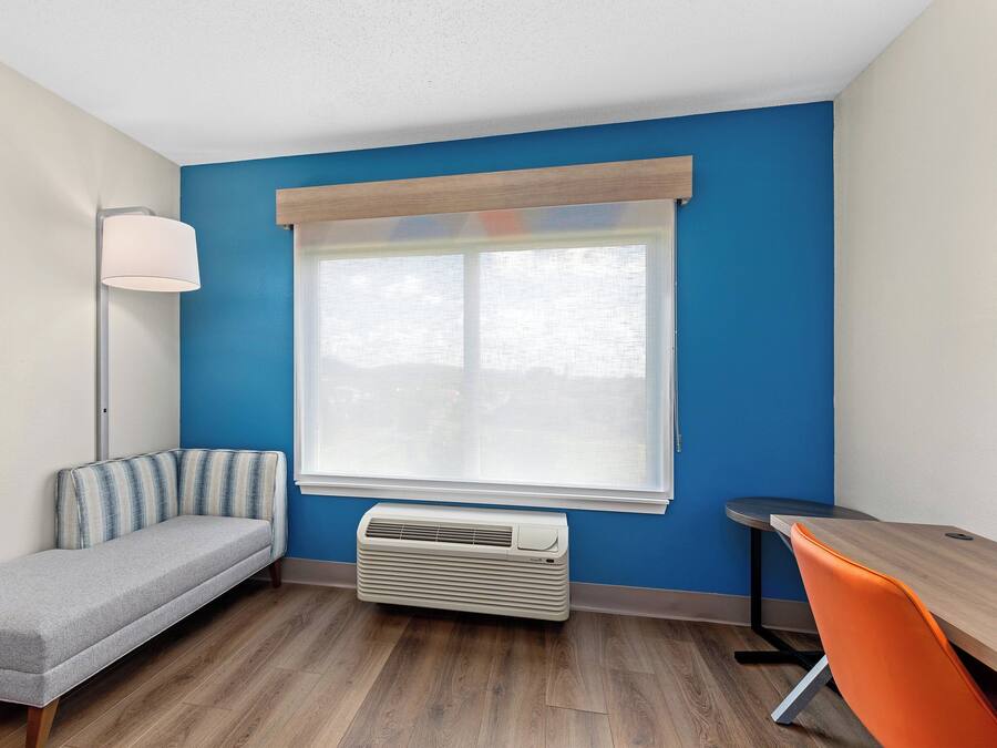 A room with a blue wall, a window with a white blind, a gray sofa, a standing lamp, a wooden table, and an orange chair.
