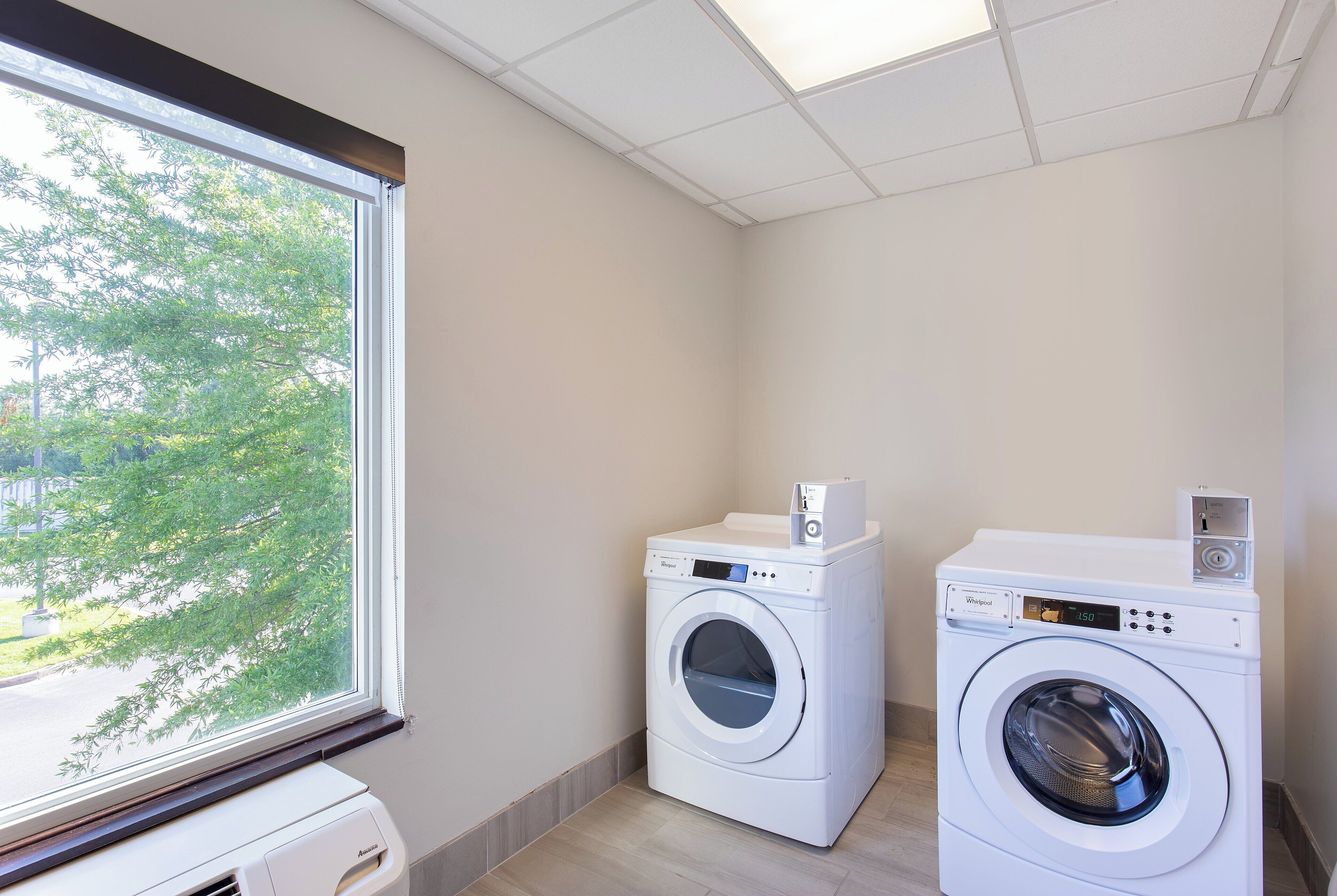 A laundry room with two front-loading washing machines and a dryer, a window with a view of trees, and a tiled ceiling.