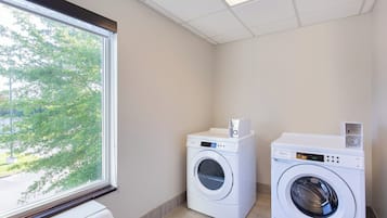 A laundry room with two front-loading washing machines and a dryer, a window with a view of trees, and a tiled ceiling.