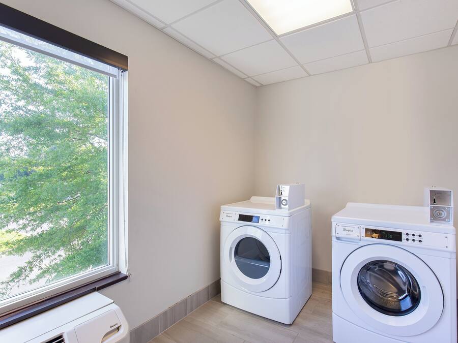A laundry room with two front-loading washing machines and a dryer, a window with a view of trees, and a tiled ceiling.
