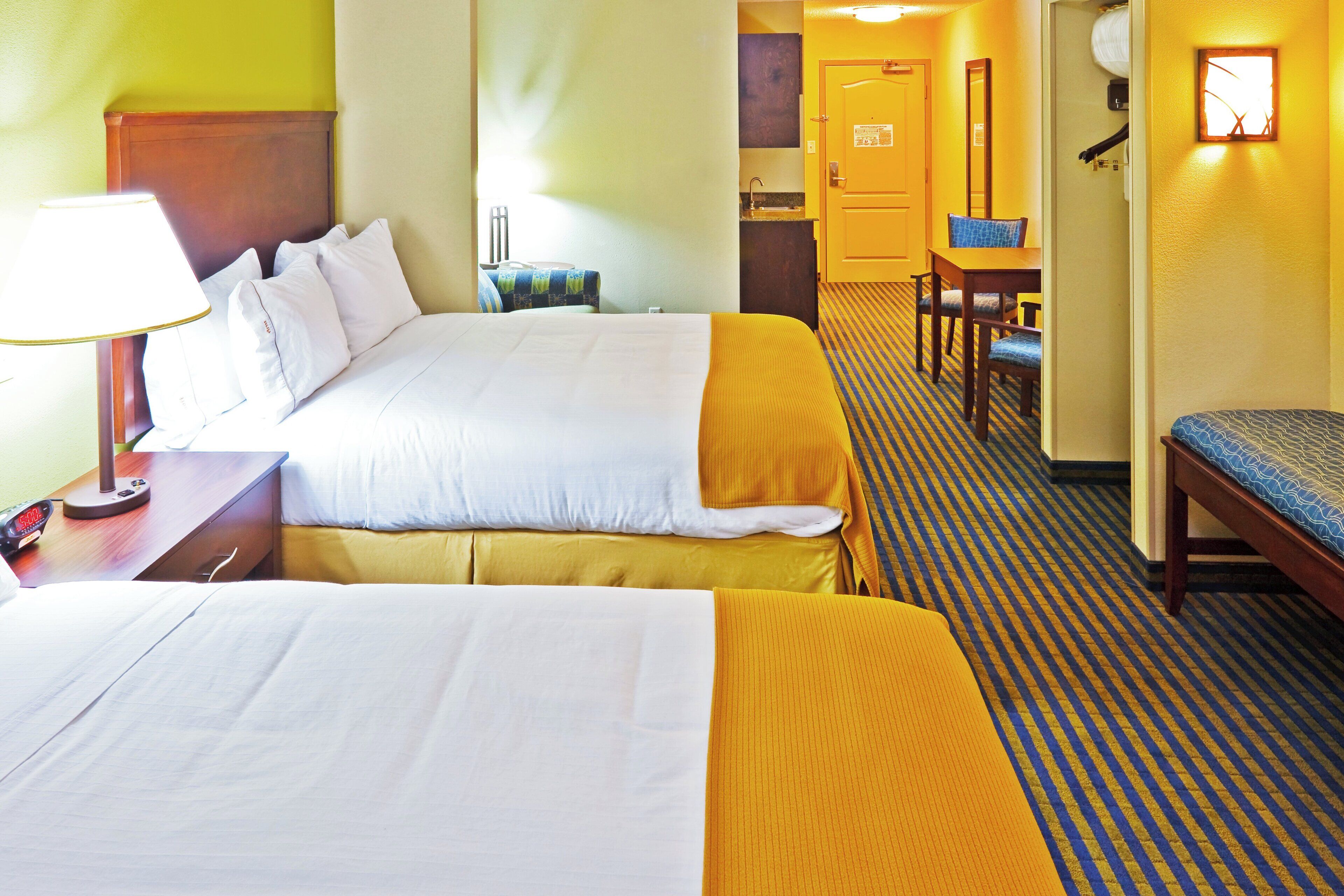 A hotel room with two beds, a desk with a lamp, a chair, and a bathroom visible in the background.