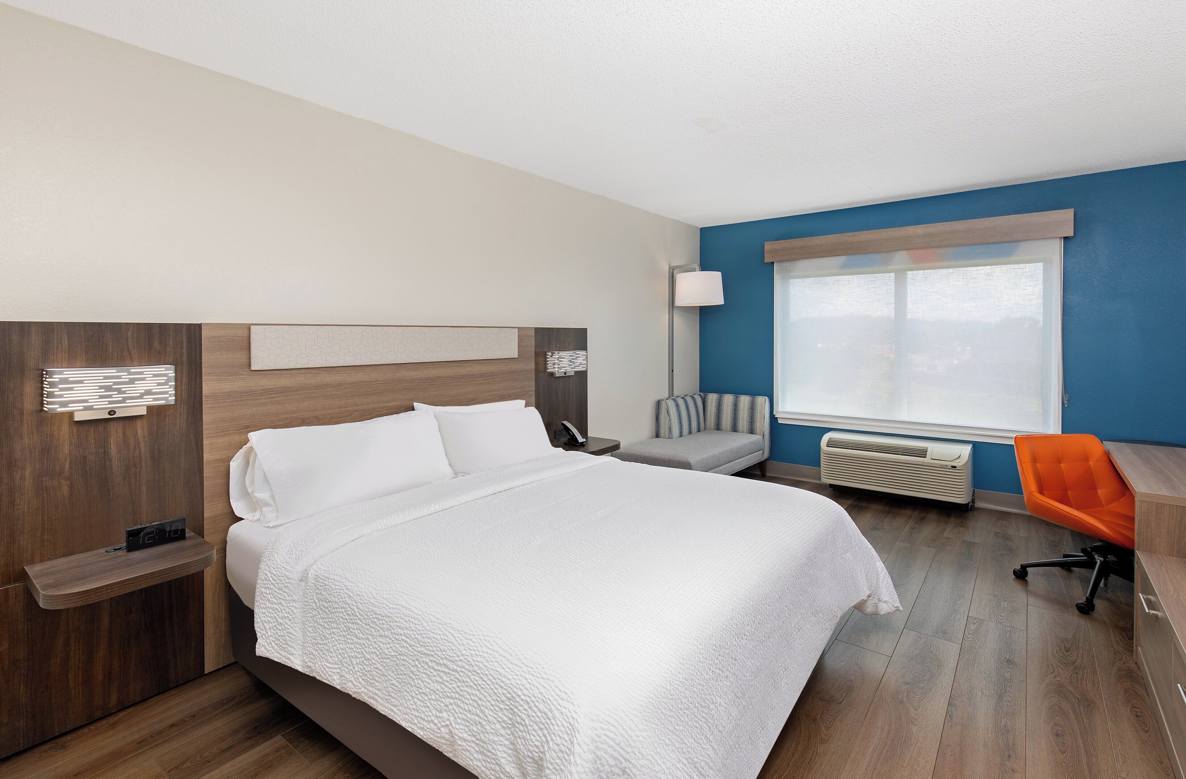 A hotel room with a large bed, a desk, an orange chair, a lamp, and a window with blinds.