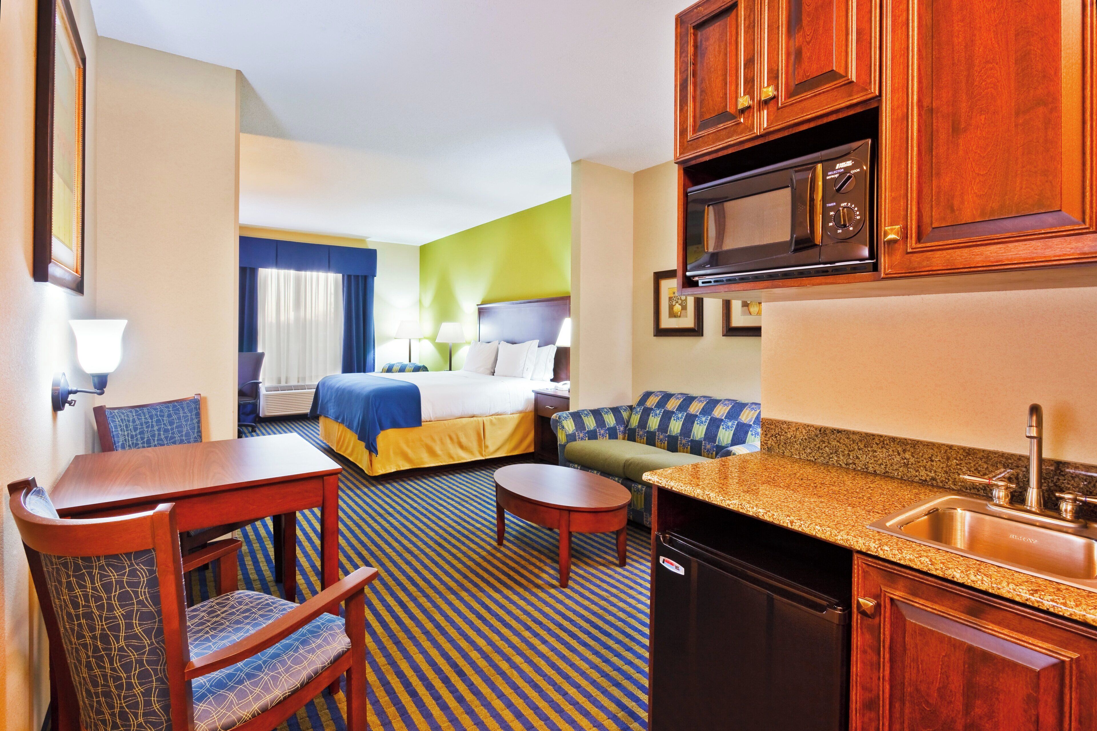 A hotel room with a bed, a sofa, a dining table, a kitchen area with a sink and stove, and a mirror.