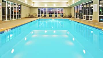 Una piscina interior con agua cristalina, rodeada de sillones de descanso y mesas, y grandes ventanales que ofrecen vistas de la ciudad de noche.