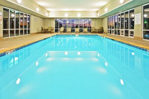 Una piscina interior con agua cristalina, rodeada de sillones de descanso y mesas, y grandes ventanales que ofrecen vistas de la ciudad de noche.