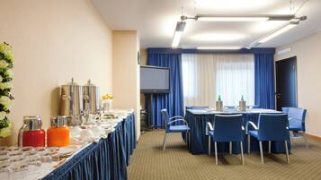 Une salle de conférence d’hôtel avec une longue table préparée pour une réunion, des chaises bleues, un écran de projection et une grande fenêtre avec des rideaux.