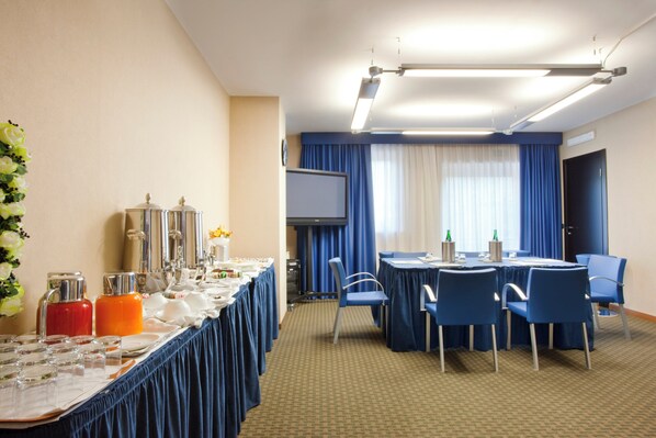Un salón de conferencias de hotel con una larga mesa dispuesta para una reunión, sillas azules, una pantalla de proyector y un gran ventanal con cortinas.