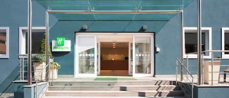 A Holiday Inn hotel entrance with a glass facade, a green awning, and a set of stairs leading up to the entrance.