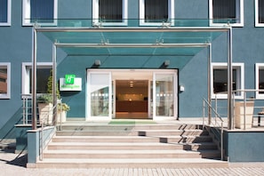 Entrada de un hotel Holiday Inn con fachada de vidrio, toldo verde y un conjunto de escaleras que conducen a la entrada.