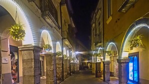Una calle nocturna con edificios de arcos, iluminada por luces colgantes y plantas suspendidas.