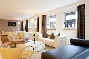 Ein modernes Wohnzimmer mit einer Couch, Sesseln, einem Couchtisch und einem Fernseher. Große Fenster mit Vorhängen, eine Lampe und eine Pflanze sind ebenfalls vorhanden.