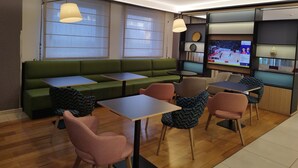 Un espace de détente moderne avec un canapé vert, plusieurs tables et des chaises. Un téléviseur fixé au mur diffuse un événement sportif.