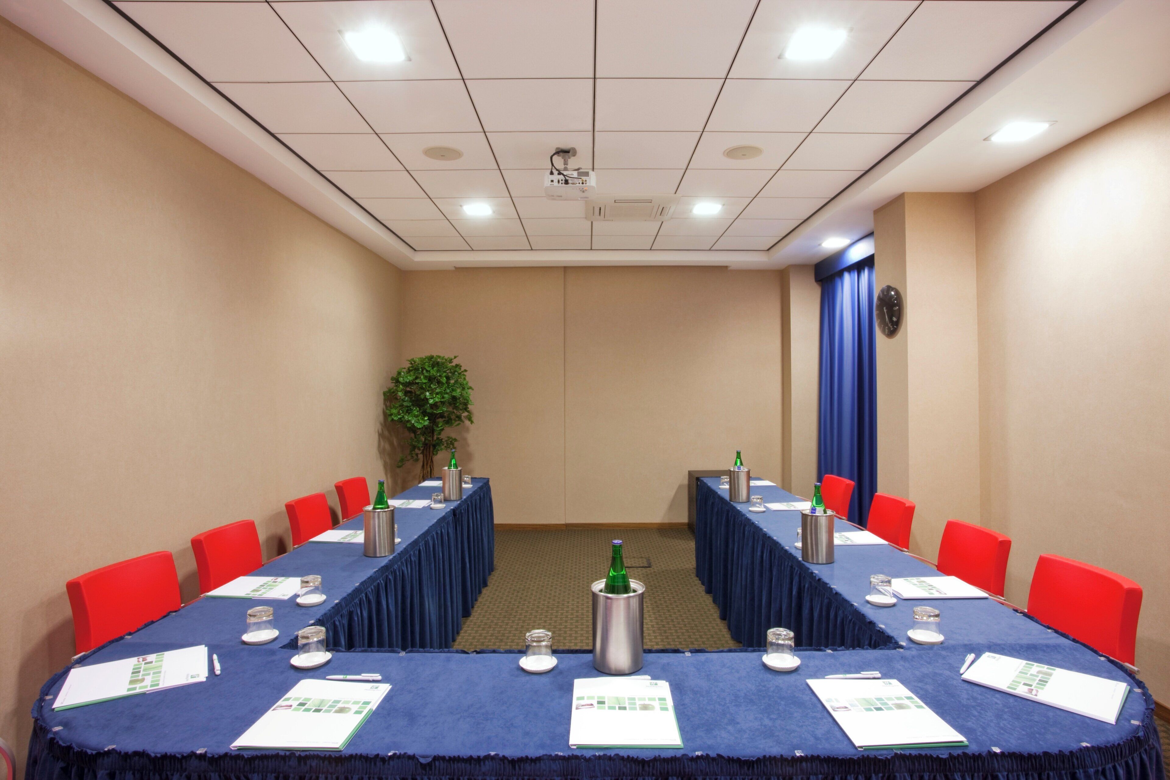 Uma sala de conferências com uma mesa comprida, cadeiras vermelhas e uma tela de projetor.
